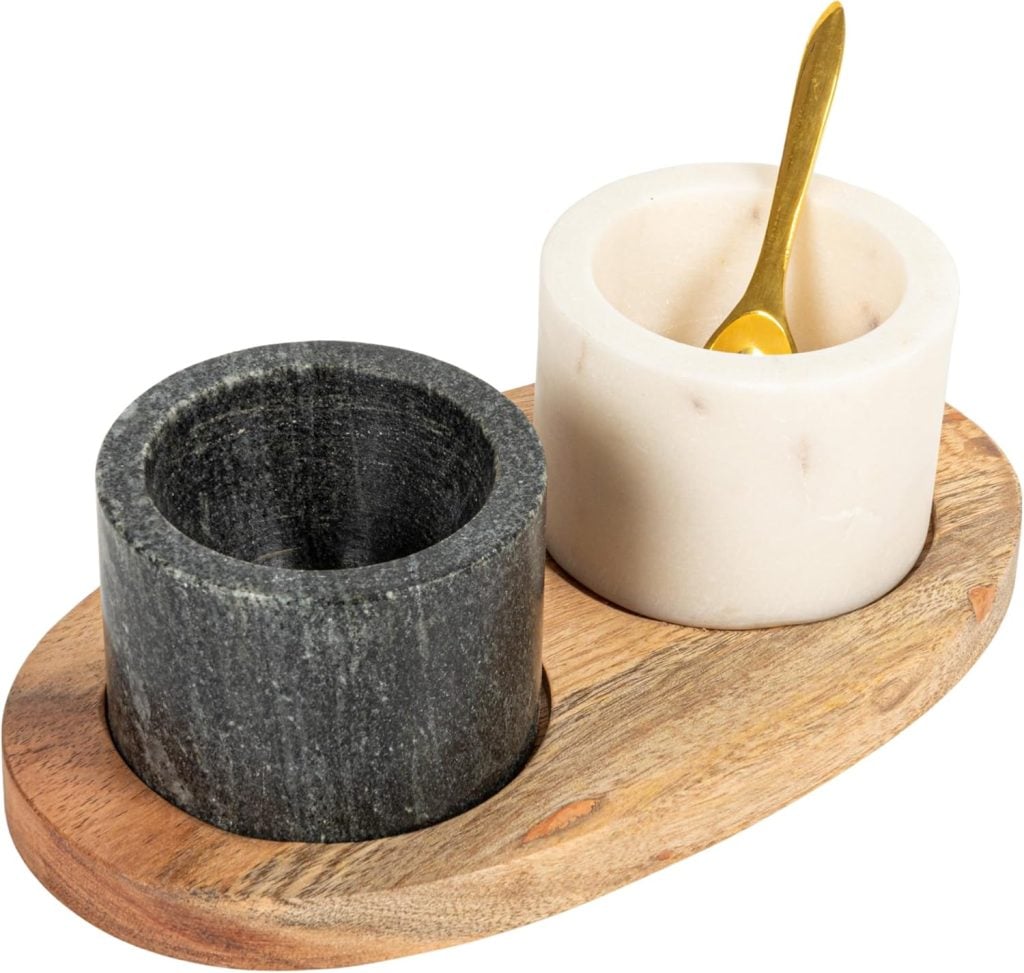Two cylindrical stone containers, one black and one white, sit on a wooden tray. The white container holds a small gold spoon.