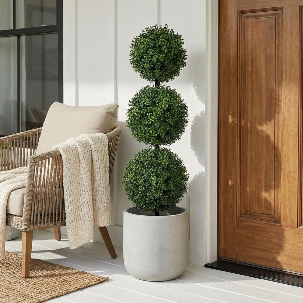 Potted topiary with three spherical layers next to a wooden door, beside a wicker chair with a beige cushion and knit throw on a white porch.