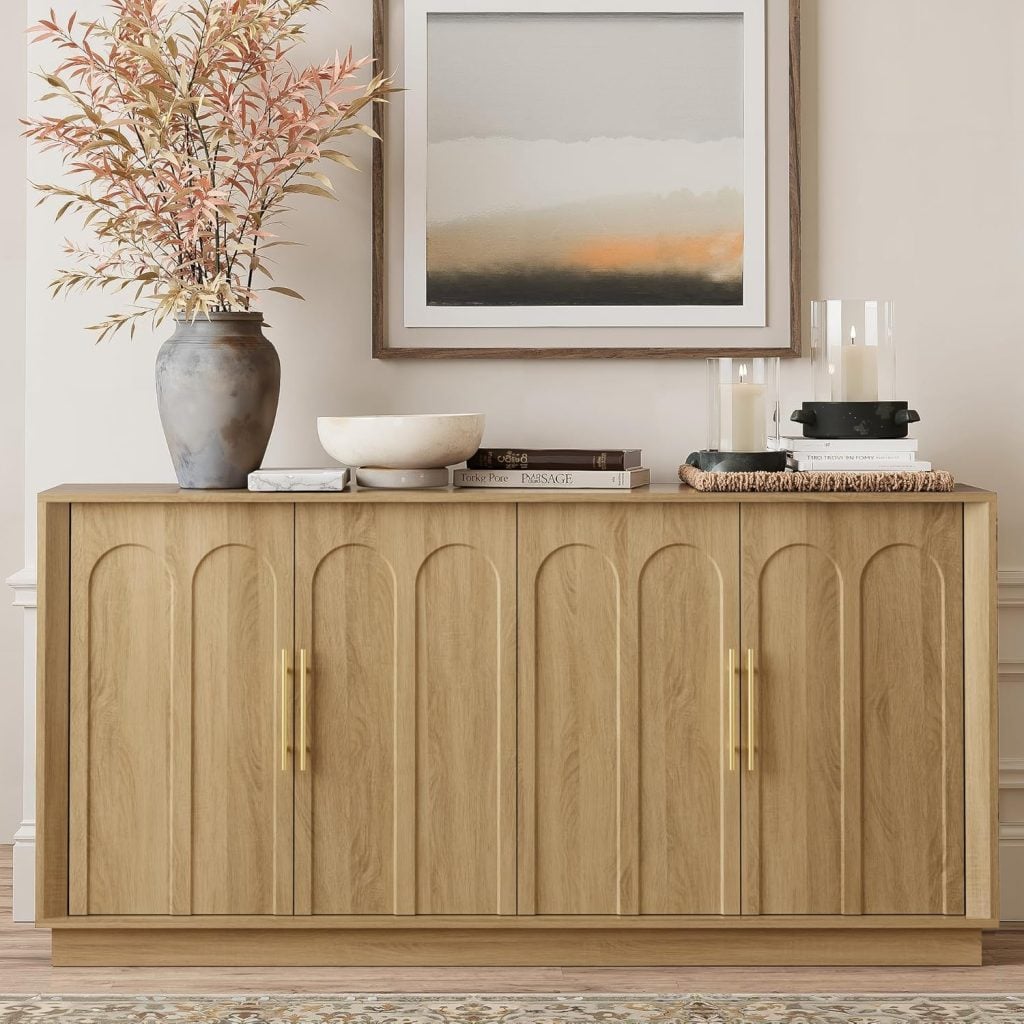 A light wood sideboard with gold handles, topped with decorative items including a vase with branches, stacked books, a bowl, and candles, positioned beneath a framed abstract painting.