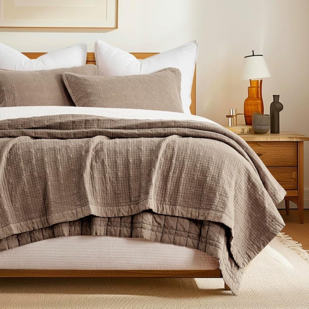 A neatly made bed with taupe bedding and white pillows stands in a softly lit bedroom next to a wooden nightstand with a lamp and decorative items.