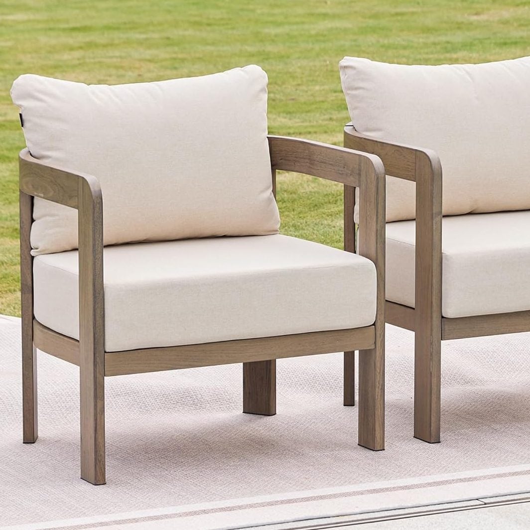 Two wooden outdoor armchairs with beige cushions, thoughtfully arranged by interior designers, sit side by side on a patio next to a swimming pool, with green grass in the background.