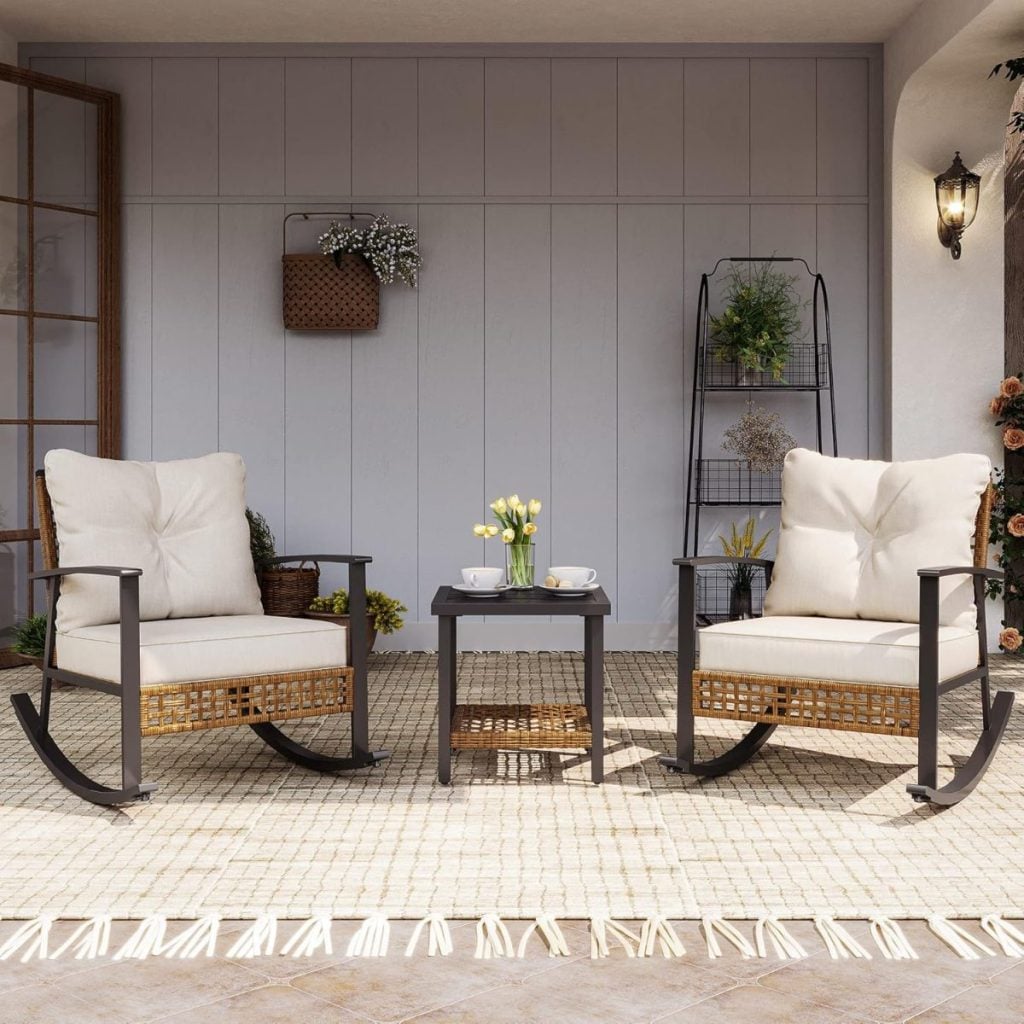 Two cushioned rocking chairs with a small table between them are arranged on a patio, with decorative plants and wall accents in the background.