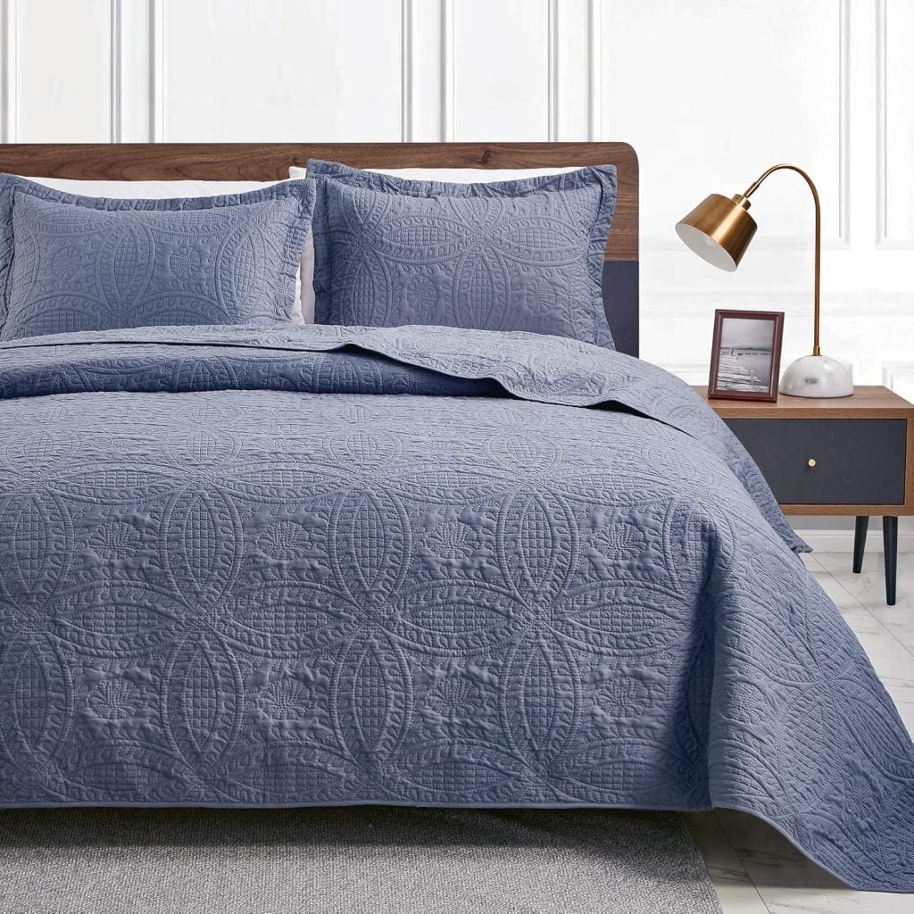 A neatly made bed with a blue quilted cover and matching pillows, next to a wooden nightstand with a lamp, picture frame, and book in a modern bedroom.