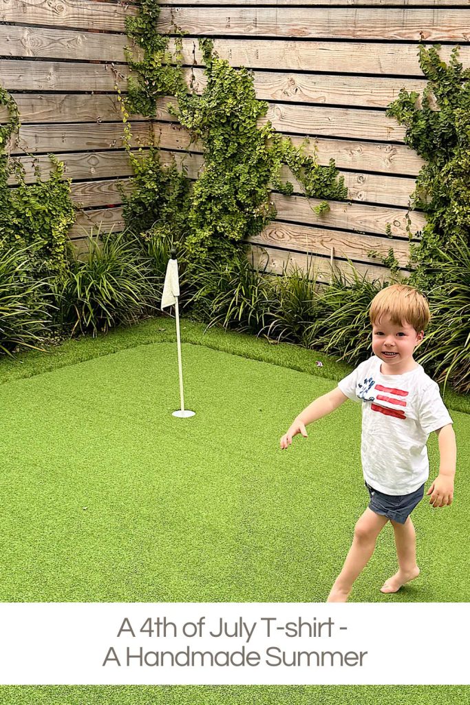A young child in a white 4th of July T-shirt walks barefoot on a small artificial grass putting green with a flag, surrounded by greenery and a wooden fence.