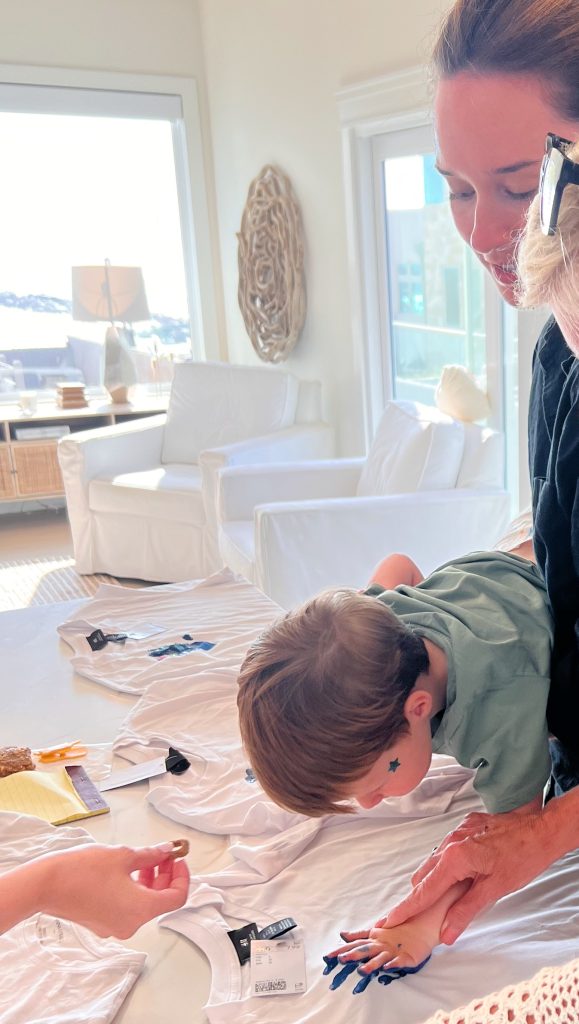 A young child is pressing their painted hand onto a white t-shirt while adults assist, inside a bright room with white furniture and large windows.
