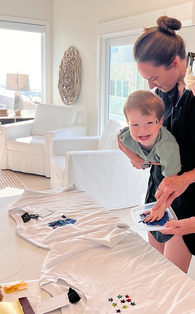 A woman helps a smiling young boy stamp colorful shapes onto a white T-shirt in a bright, sunlit room.
