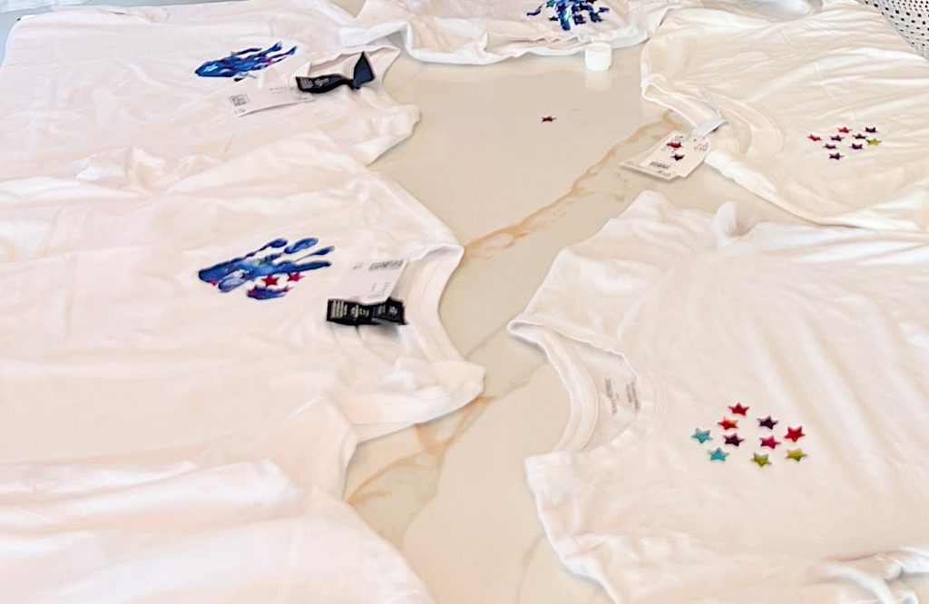 Six white t-shirts are spread out on a table, each decorated with star-shaped stickers or painted handprints in various colors.