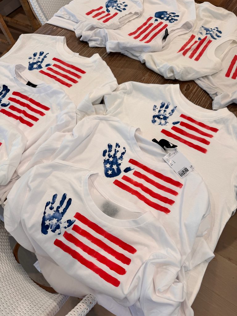 Several white t-shirts with handprint stars and red stripes form stylized American flags, arranged on a wooden table.