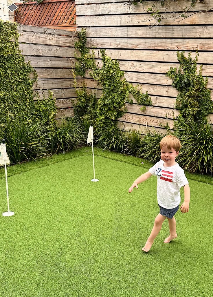 A young child walks barefoot on artificial grass in a backyard, with two small golf flags and greenery along a wooden fence.