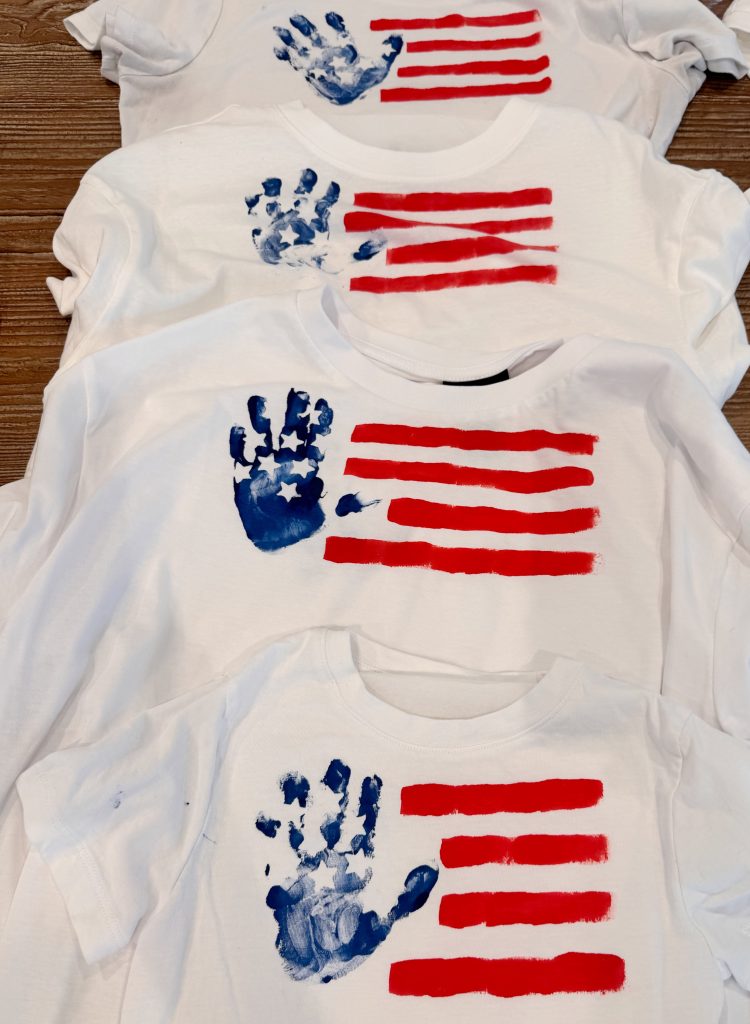 Four white t-shirts with painted American flags, featuring red stripes and a blue handprint in place of the stars, are laid out on a wooden surface.