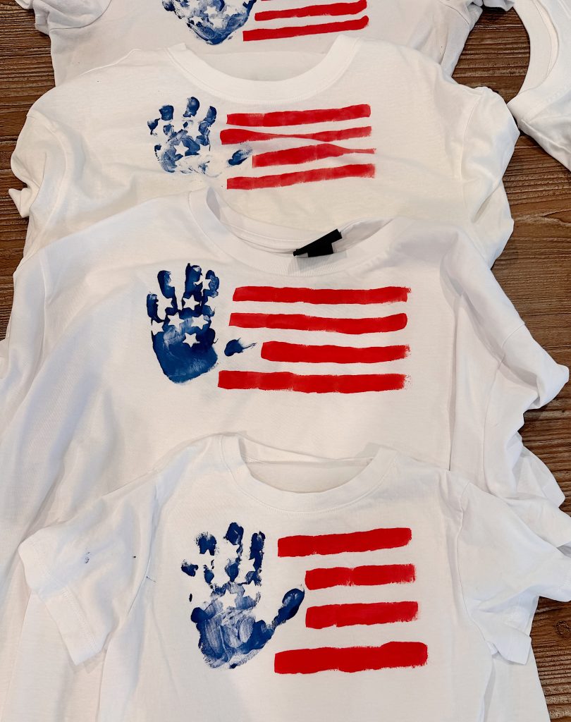 Four white shirts with American flag designs made from red painted stripes and blue handprints for the stars, laid out on a wooden surface.