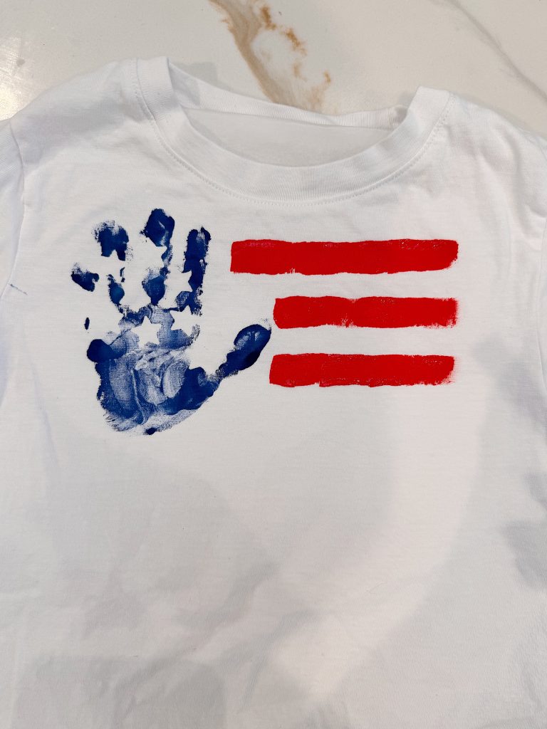 White t-shirt featuring a blue handprint and three red painted stripes, resembling a stylized version of the American flag.