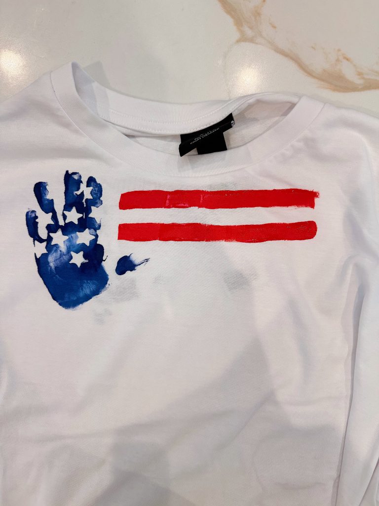 White shirt featuring a blue handprint with white stars and two horizontal red stripes, resembling an abstract American flag design.
