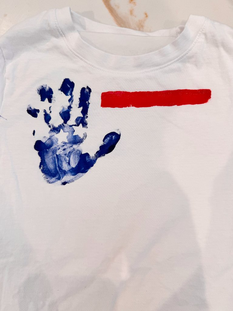 A white shirt with a blue handprint and white stars resembling the U.S. flag, next to a horizontal red stripe painted above it.