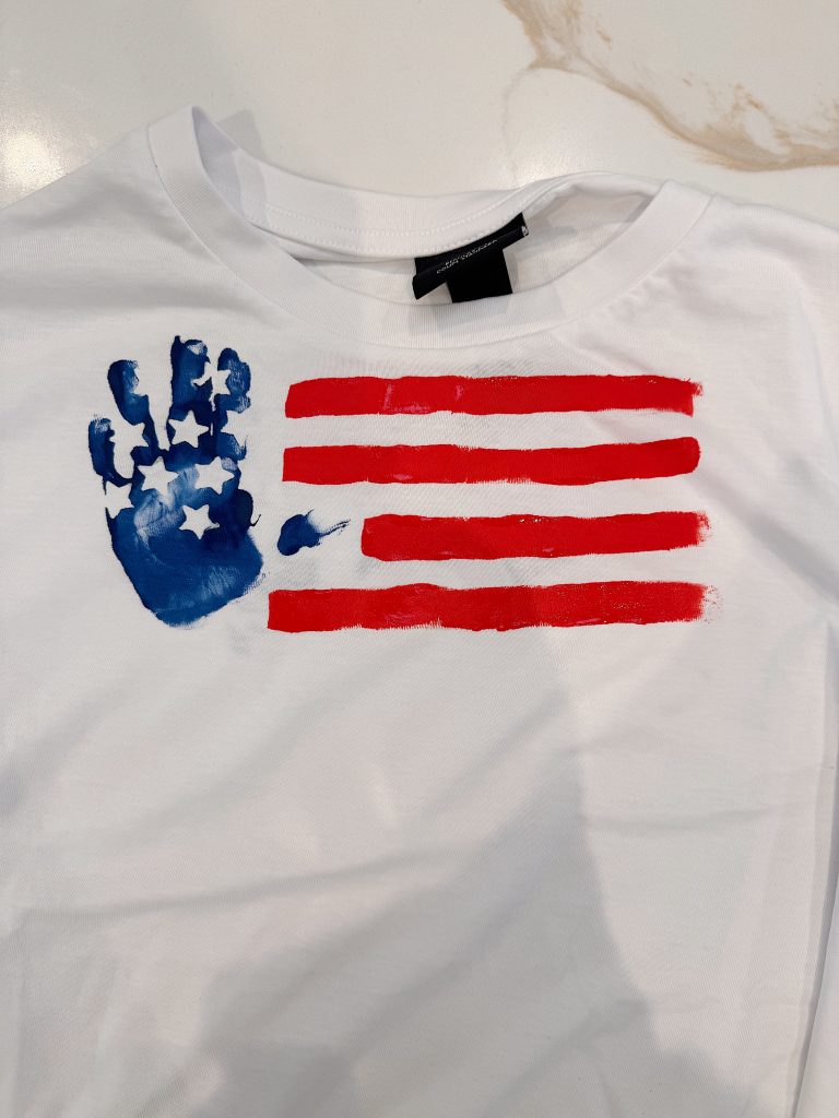 White T-shirt featuring a handprint in blue with white stars next to red painted stripes, resembling the American flag.