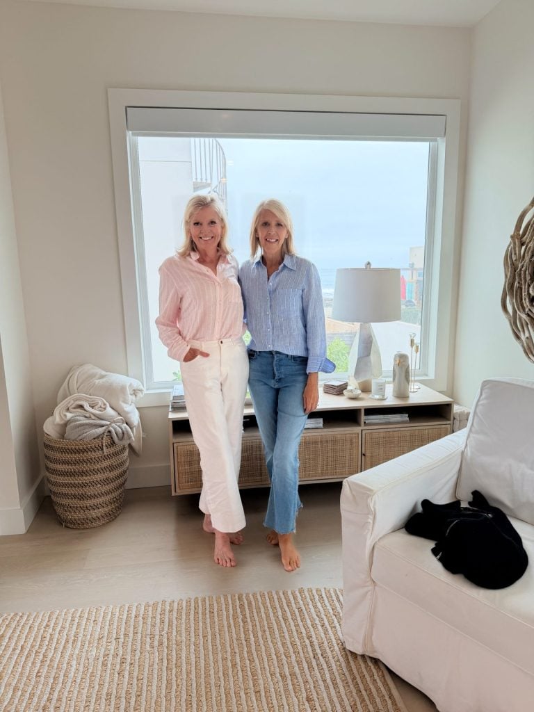 Two women stand barefoot in front of a large window in a bright, modern living room with neutral decor and a view of the outdoors.