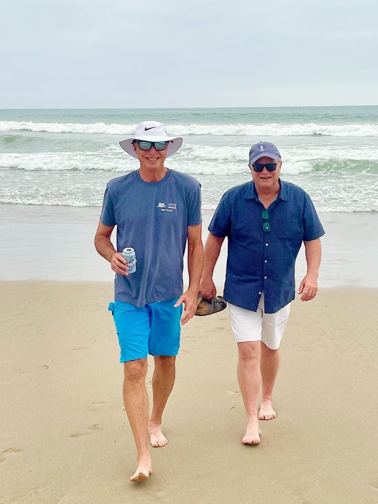 Two men walk barefoot on a sandy beach near the ocean, both wearing sunglasses and hats. One holds a drink can; the other carries sandals. The sky is overcast.