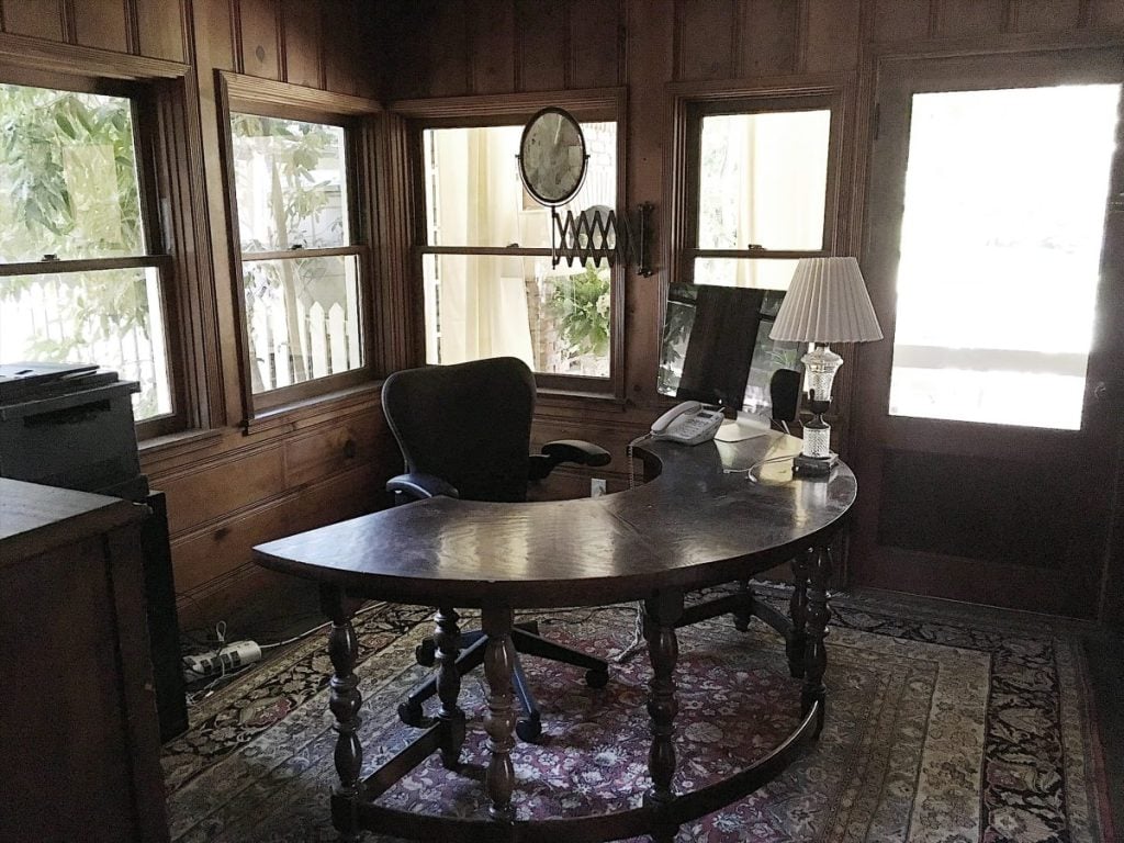 A home office with a curved wooden desk, office chair, desktop computer, desk lamp, telephone, and clock on a wood-paneled wall, with windows and a door letting in natural light.