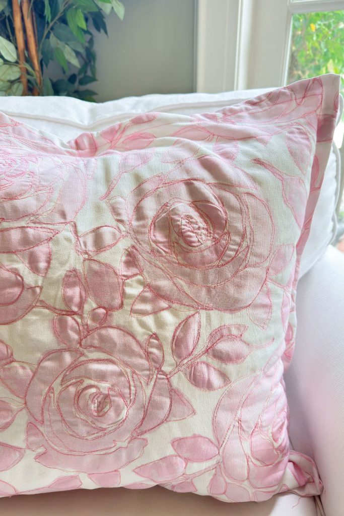 A white pillow with pink embroidered floral patterns rests on a white sofa near a window and green plant.