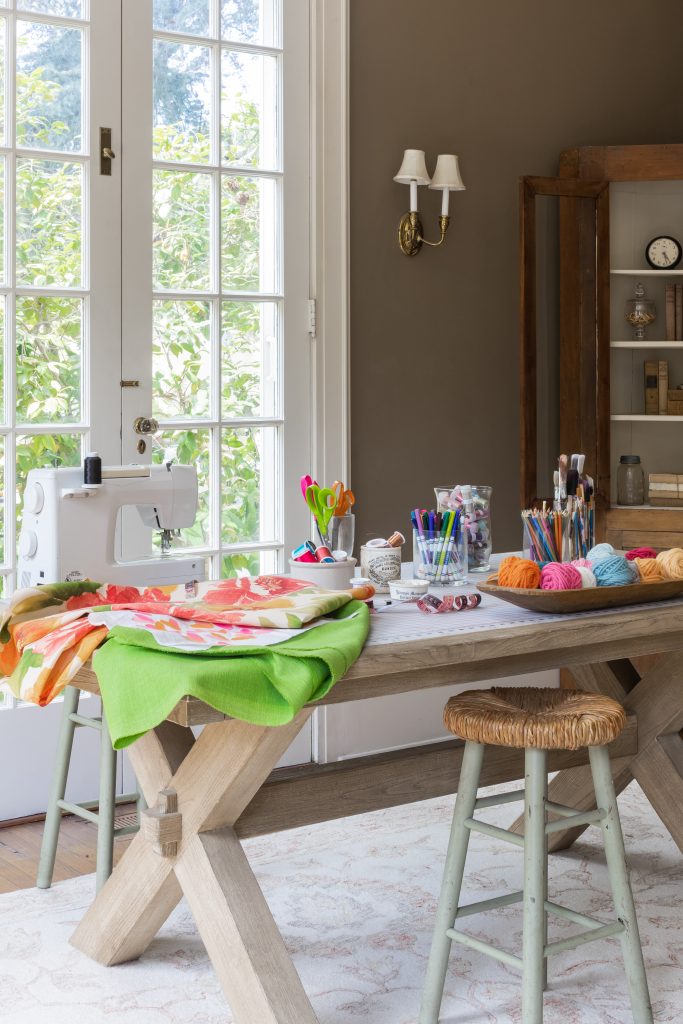 A craft room with a large wooden table holding a sewing machine, art supplies, yarn, scissors, and fabric, next to tall windows with garden views.
