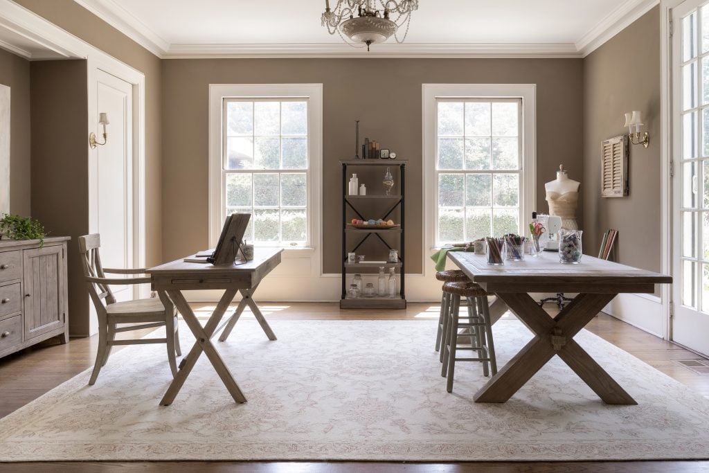 A bright room with two wooden tables, chairs, a central shelving unit, and large windows letting in natural light, furnished in a neutral color palette.