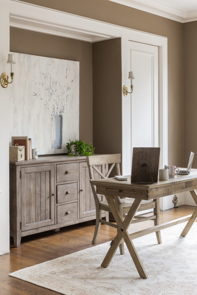 A home office with a wooden desk and chair, a laptop, a sideboard with a potted plant, a large floral painting, and neutral brown walls.