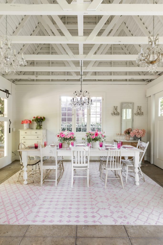 A white dining room with a large table, six chairs, pink floral arrangements, chandeliers, a patterned rug, and exposed beams on the ceiling.