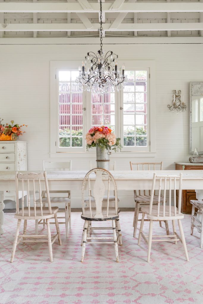 A dining room with a white wooden table, mismatched vintage chairs, a metal vase with flowers, a crystal chandelier, and large windows letting in natural light.