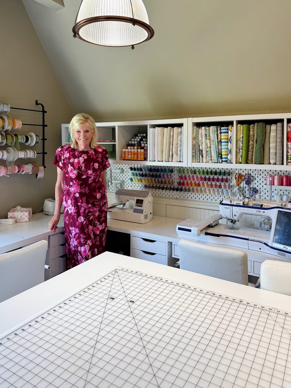A woman in a pink floral dress stands in a well-organized sewing room with fabric, thread, ribbon, and sewing machines neatly arranged on shelves and tables.