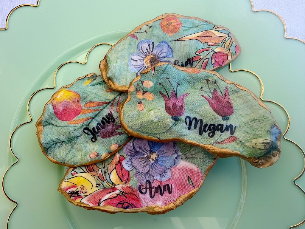 Three oyster shells with colorful floral designs and handwritten names (Jenny, Megan, Ann) are arranged on a green plate with a scalloped gold rim.