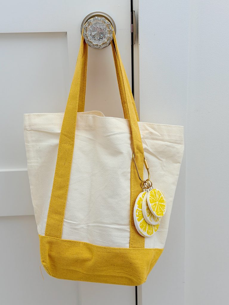 A beige and yellow canvas tote bag hangs on a glass doorknob, with two round keychains featuring yellow citrus slice designs attached to the handle.