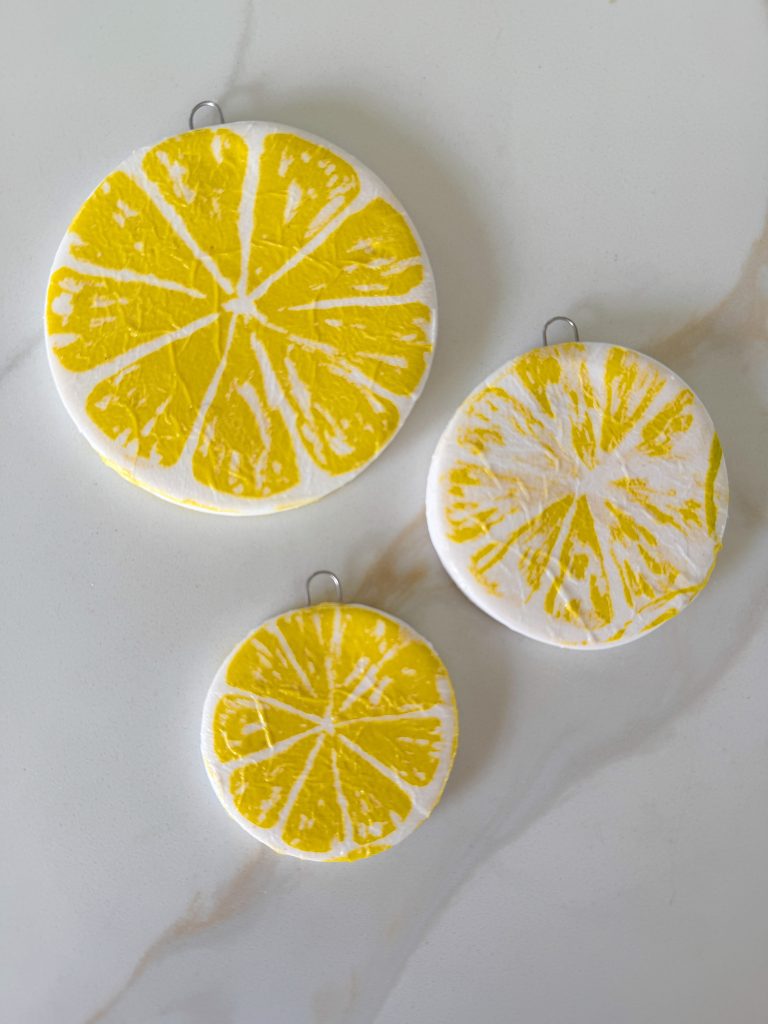 Three round ornaments with yellow citrus slice designs and metal hooks are arranged on a light marble surface.