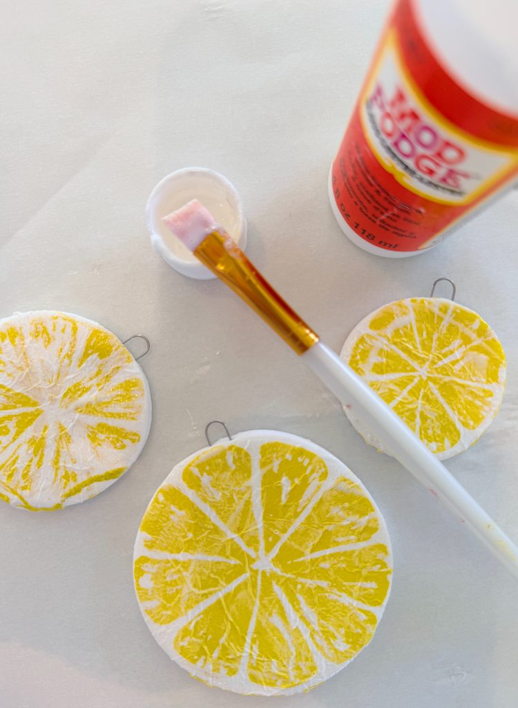 Round ornaments painted with yellow citrus slices are being coated with Mod Podge using a paintbrush; the bottle and brush are visible on a white surface.