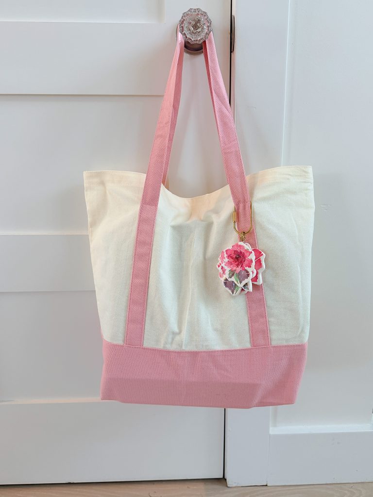 A cream and pink tote bag with pink handles hangs on a door knob. A flower-shaped keychain is attached to one handle.