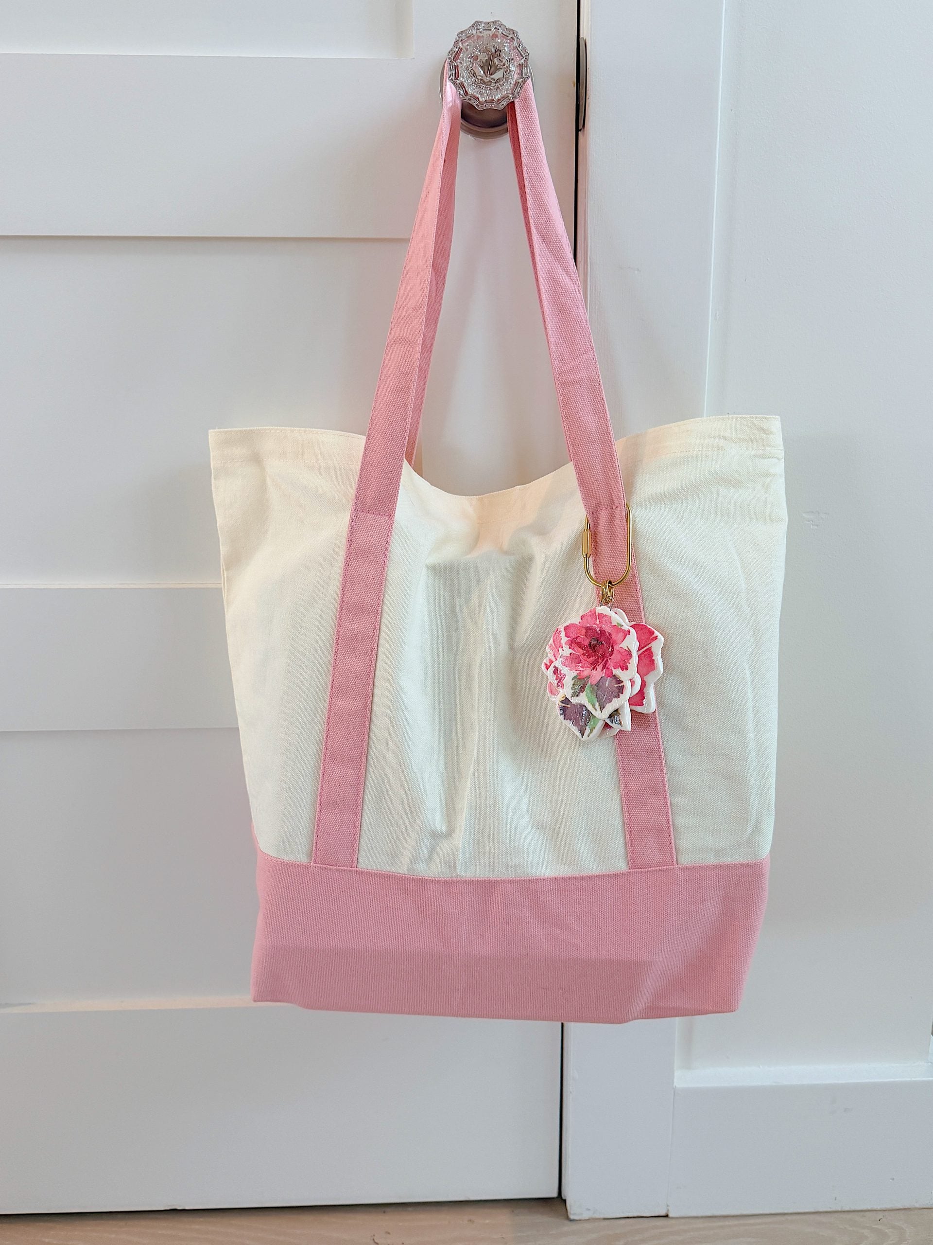 A cream and pink tote bag with pink handles hangs on a door knob. A flower-shaped keychain is attached to one handle.