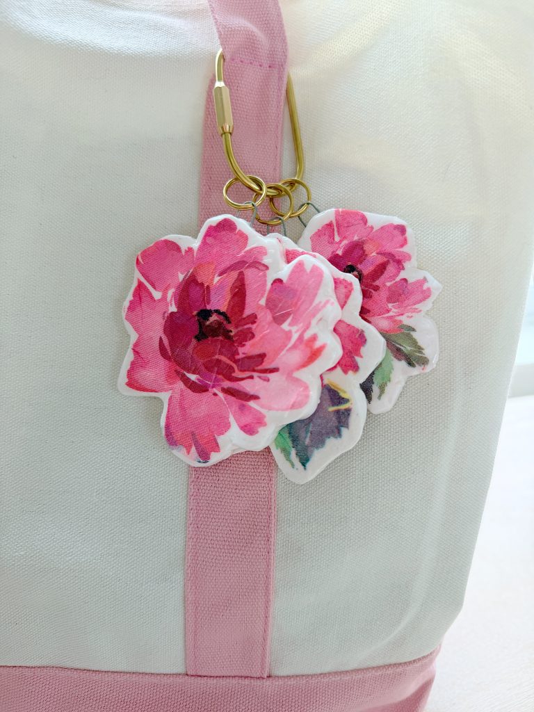 Two pink watercolor flower keychains hang from a gold carabiner attached to a white and pink fabric bag.