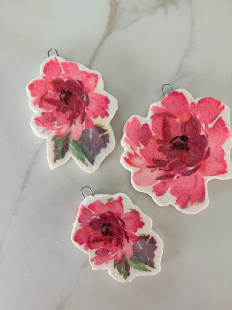 Three handmade ornaments shaped like red and pink flowers with green leaves, lying on a light marble surface.