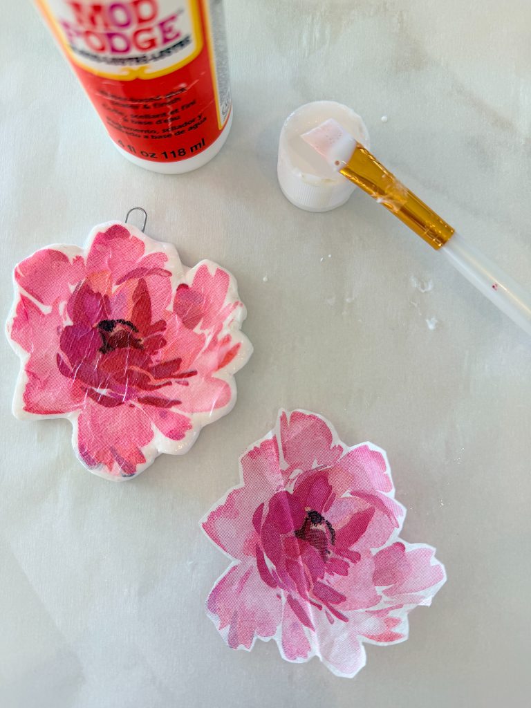 Two pink floral cut-outs, a paintbrush with glue residue, and a bottle of Mod Podge on a light surface, suggesting a crafting project in progress.