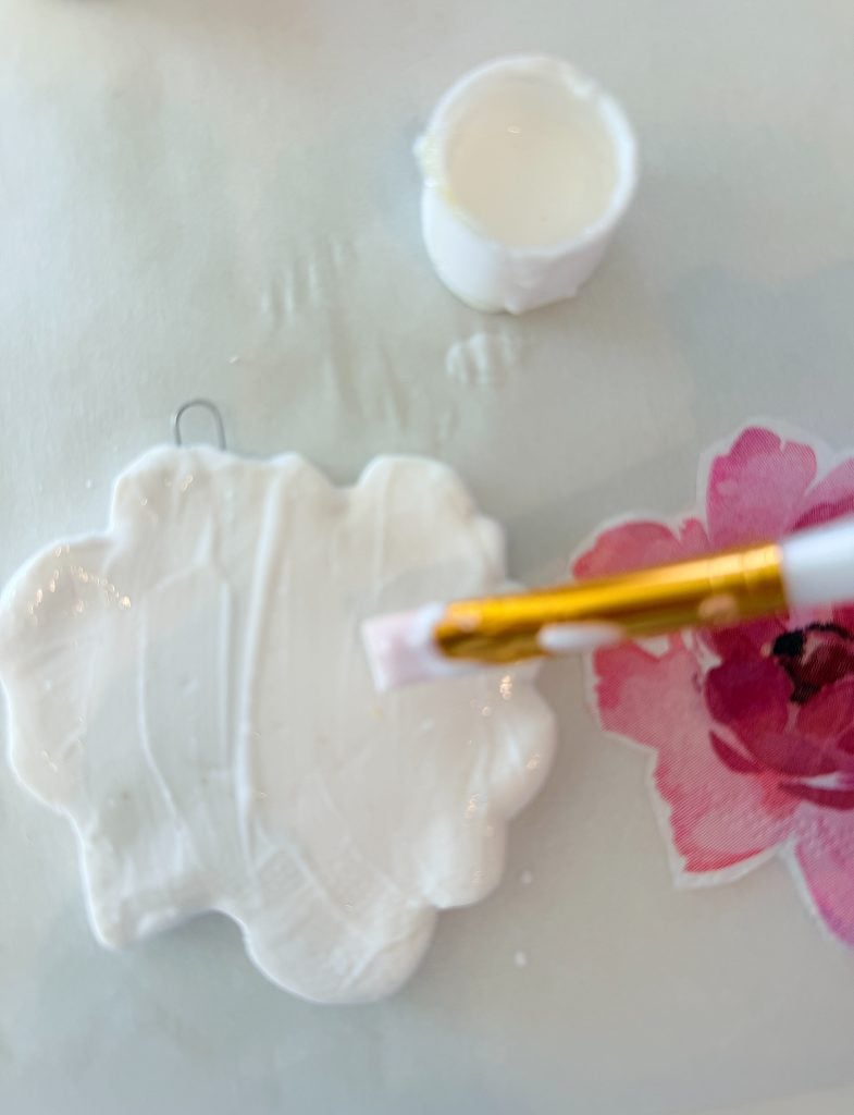 A paintbrush applies white paint to a cloud-shaped object near a white cap and a pink flower graphic on a light surface.