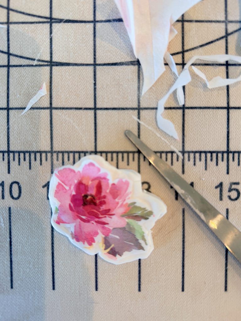 A pair of scissors next to a floral sticker and paper scraps on a gridded cutting mat.