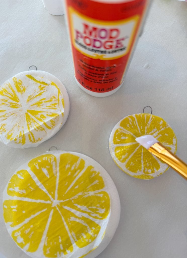 A paintbrush applies Mod Podge to round ornaments painted with yellow lemon slice designs on a white surface.