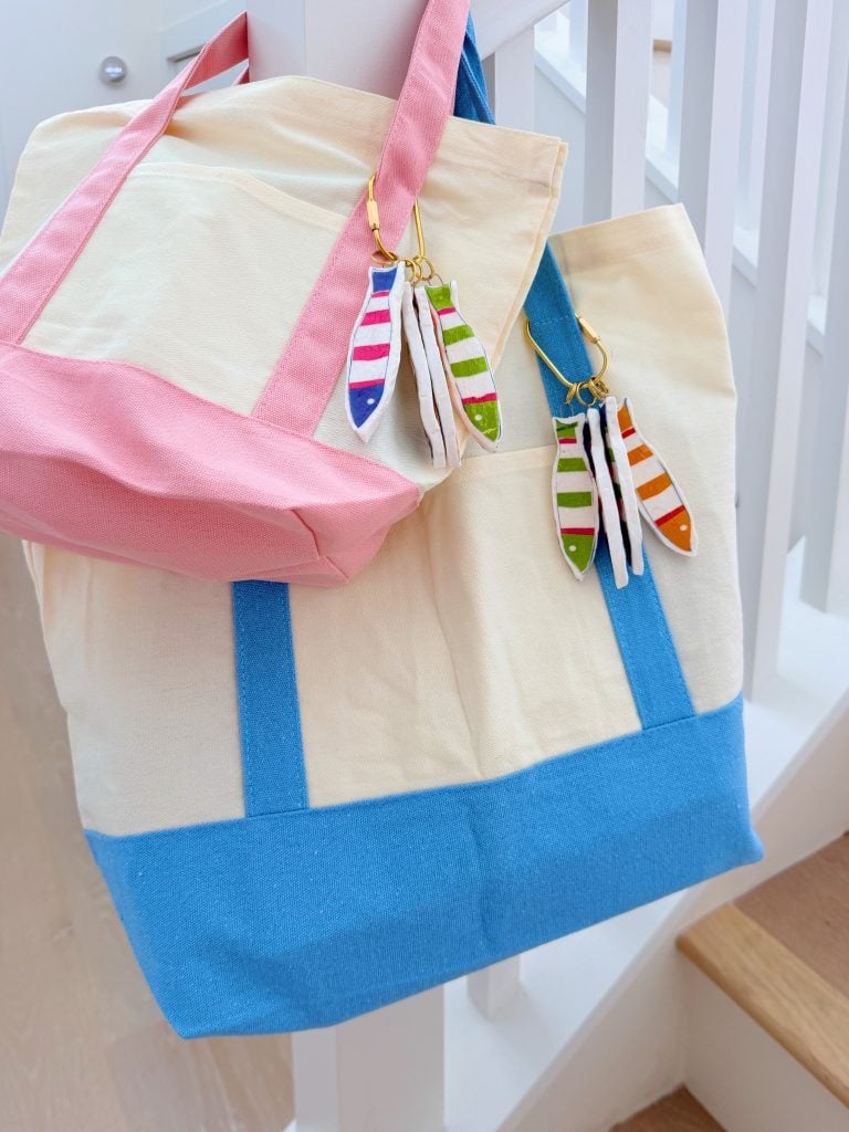 Two canvas tote bags, one with pink straps and one with blue straps, hang on a staircase railing. Both have colorful feather-shaped keychains attached.