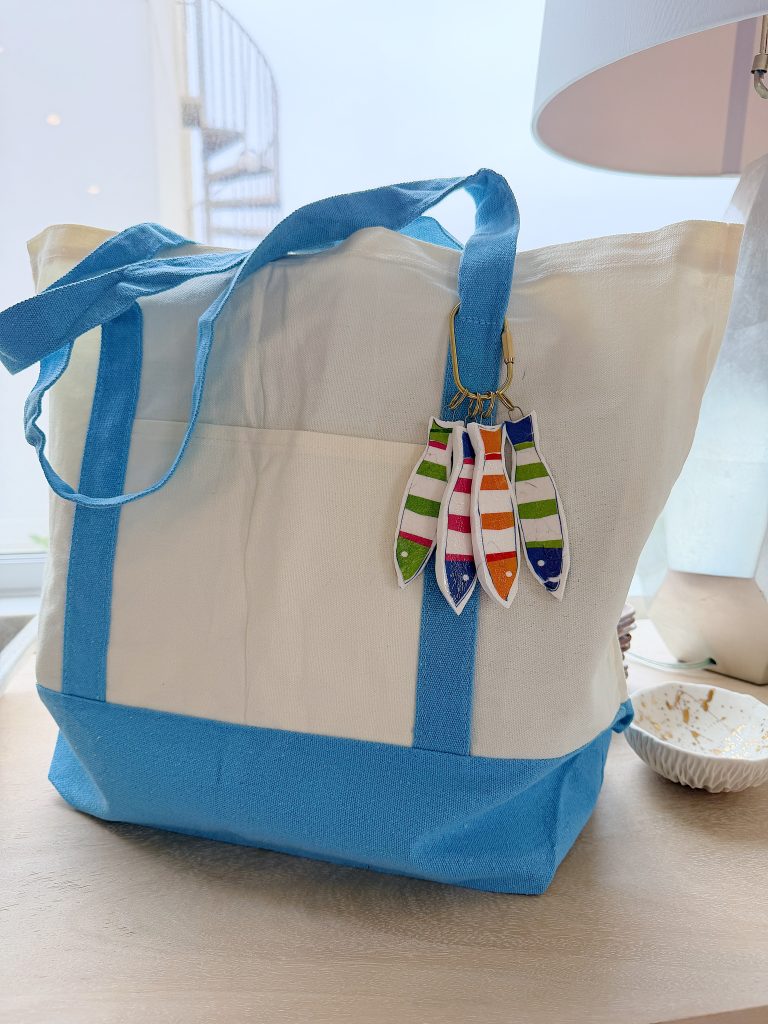 A blue and white tote bag with striped fish-shaped keychains hangs on a table next to a white lamp and a small dish.