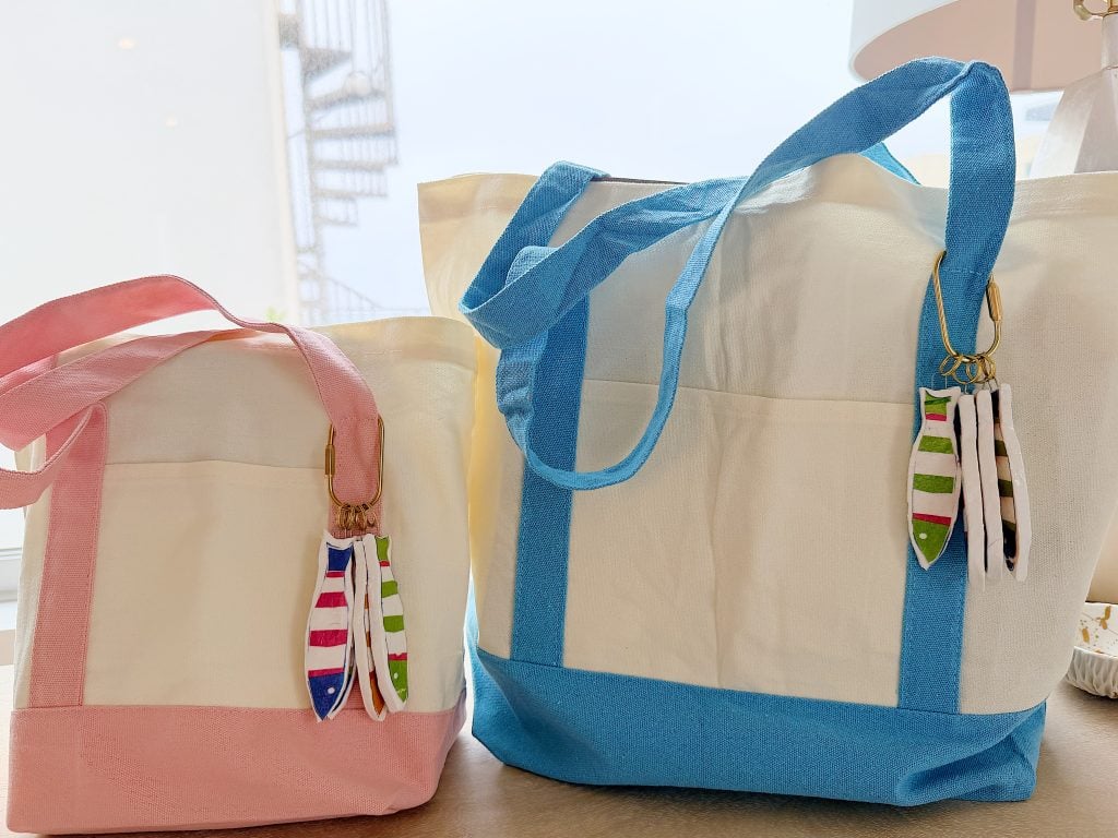 Two canvas tote bags, one with pink handles and one with blue handles, each decorated with striped fish-shaped keychains, placed on a table indoors.