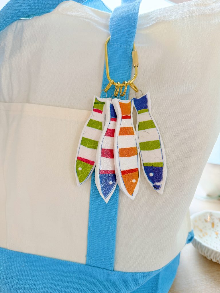 A white and blue bag with a gold keyring holding four striped fish-shaped keychains in green, pink, orange, and blue.