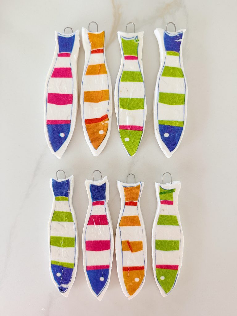 Seven fish-shaped decorations with colorful horizontal stripes in blue, orange, green, and pink are arranged on a light surface. Each has a wire loop at the top.