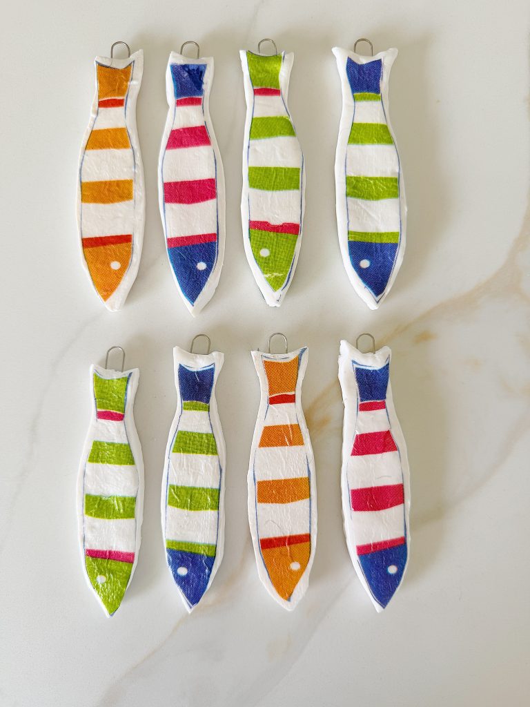 Eight fish-shaped ornaments with colorful horizontal stripes in orange, red, green, and blue, arranged in two rows on a light surface.