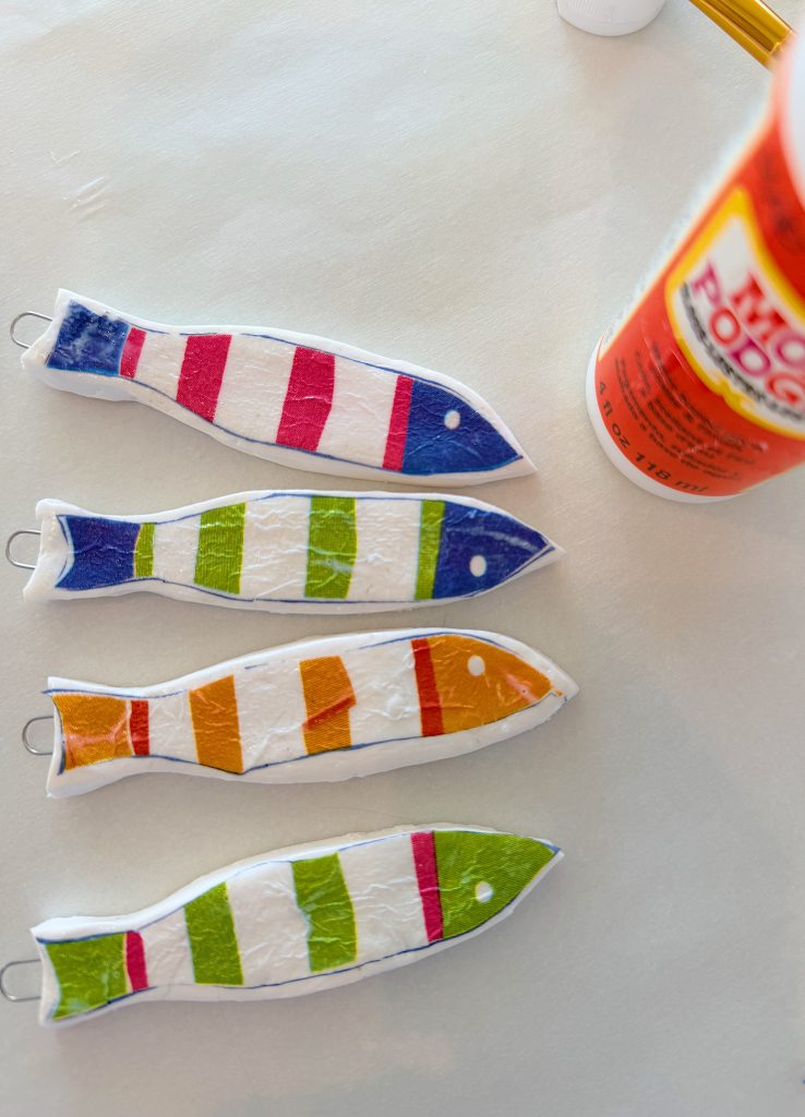 Four colorful fish-shaped crafts with striped patterns are laid out on a table next to a bottle of Mod Podge glue.
