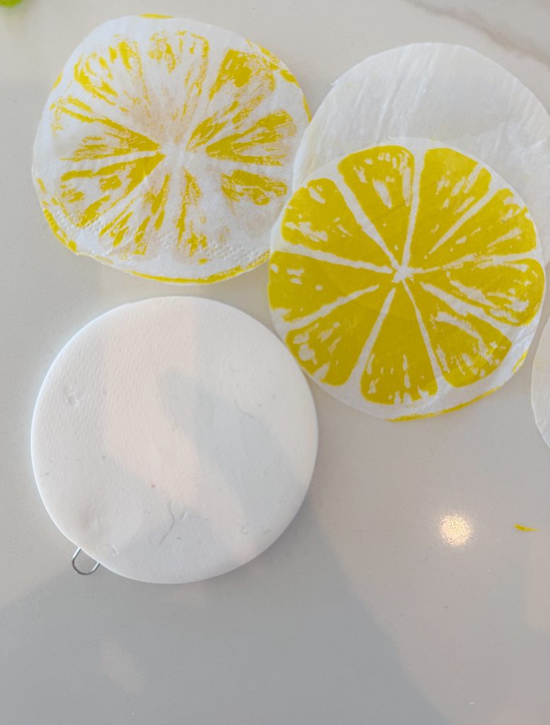 Three round pieces of thin paper with yellow citrus designs sit on a white surface next to a plain white round disk with a metal loop.