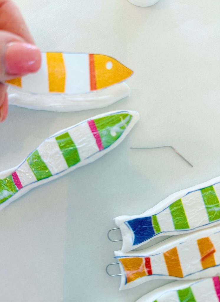 A hand holds a striped fish-shaped craft, with several similar colorful fish-shaped pieces and metal hooks on a white surface.